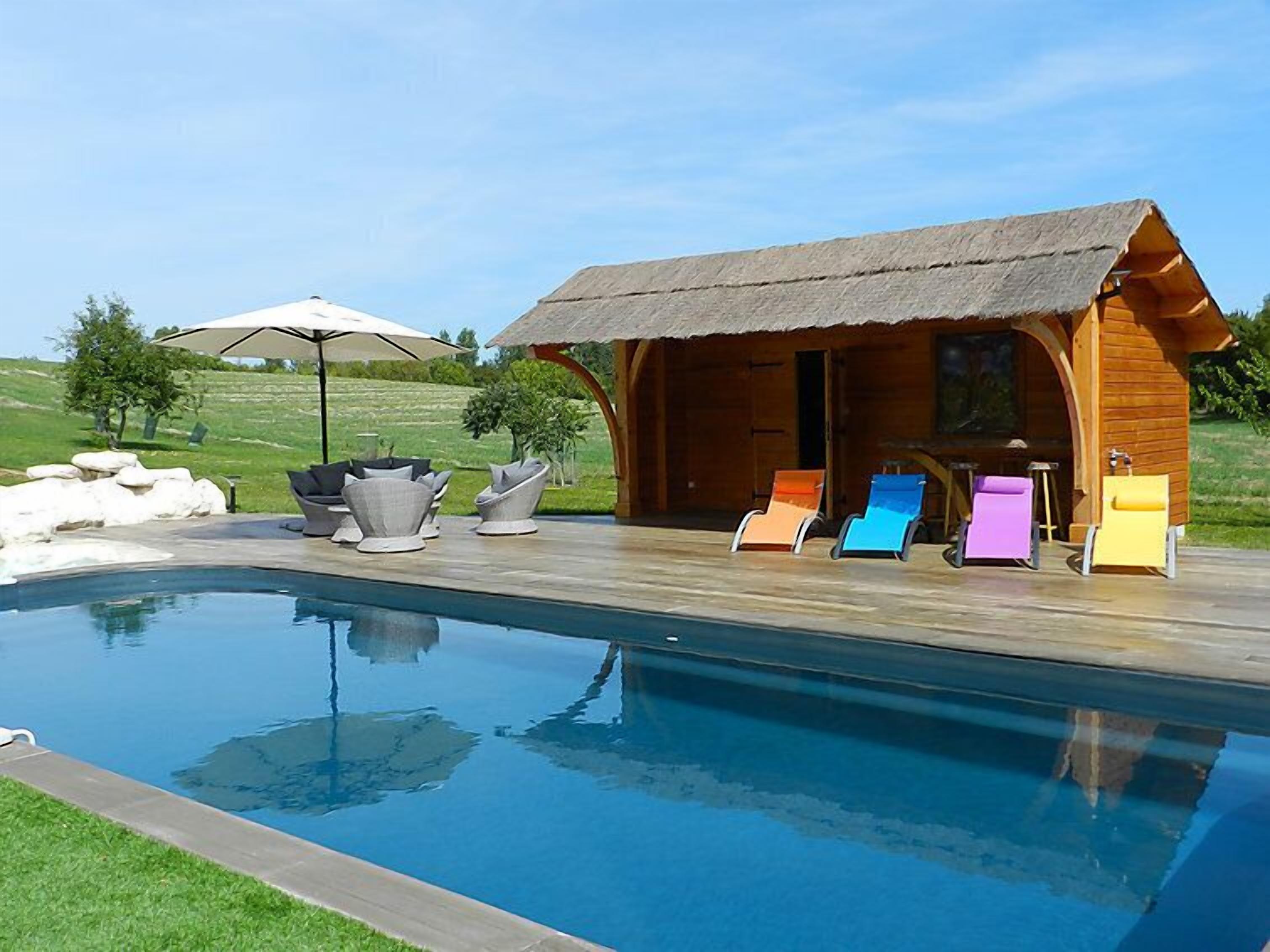 A wooden cabin with a thatched roof, a swimming pool, and colorful lounge chairs.