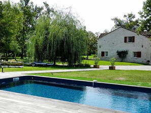 Une piscine entourée d’une terrasse en pierre, un bâtiment en pierre avec des volets rouges et un saule pleureur.