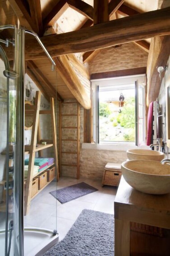 A rustic bathroom with wooden beams, a glass shower enclosure, and a large window offering a view of greenery.