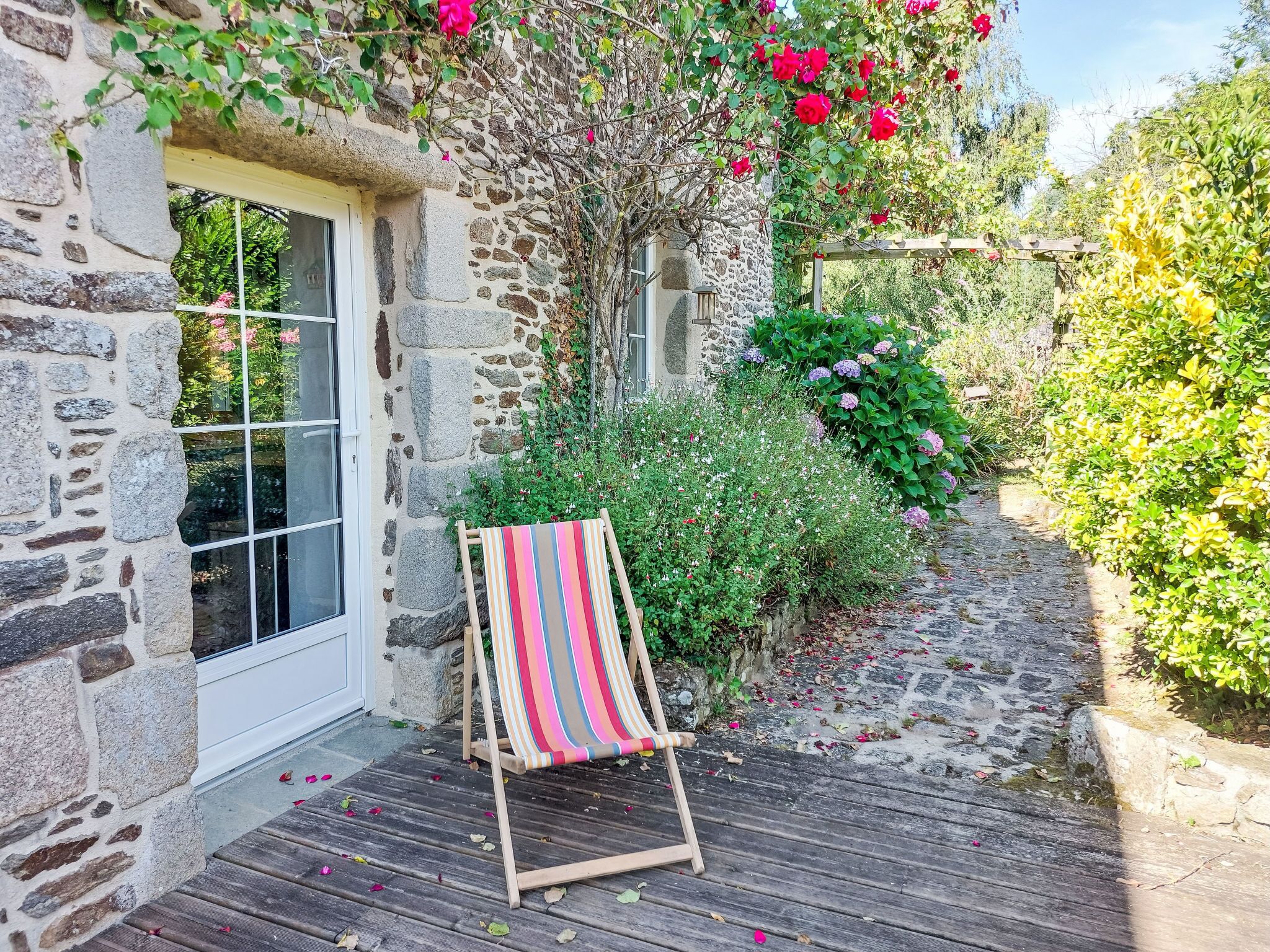A stone building with a wooden deck, a striped beach chair, and lush greenery including roses and hydrangeas.