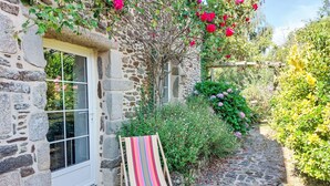 A stone building with a wooden deck, a striped beach chair, and lush greenery including roses and hydrangeas.