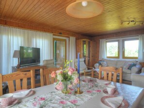 Ein gemütliches Blockhaus-Interieur mit Holzbalkendecke, einem Fernseher auf einem Holzbodenständchen, einem Esstisch mit floralem Tischläufer und einer Couch mit Kissen.