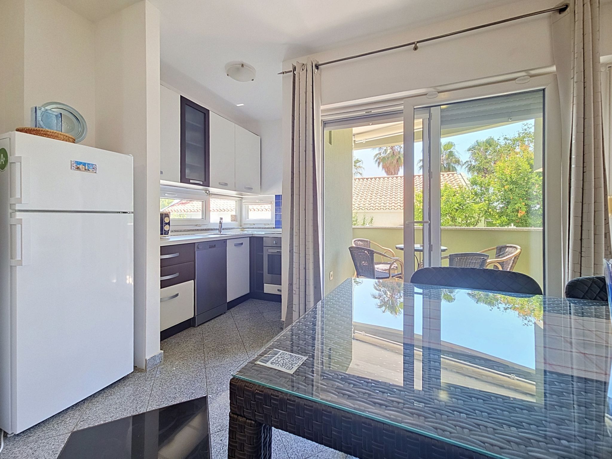 A modern kitchen with a refrigerator, microwave, and cabinets, leading to a balcony with a glass table and chairs.