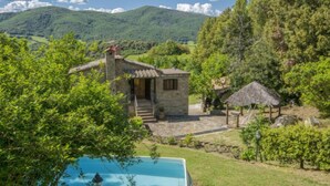 Une maison en pierre avec une petite piscine, entourée d’une végétation luxuriante et avec des montagnes en toile de fond.