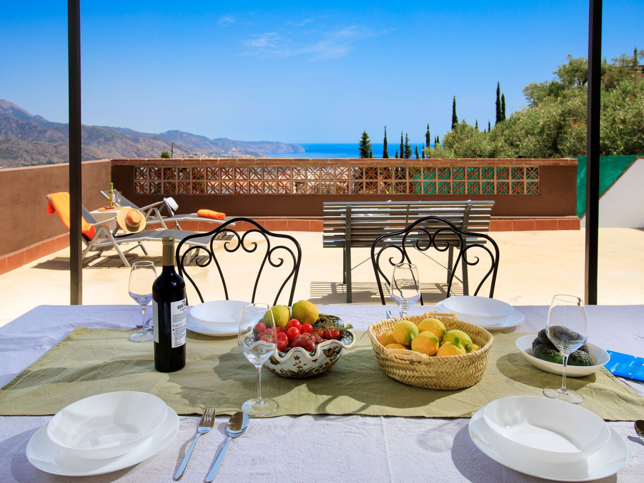 A table set for two with a bottle of wine, glasses, and a fruit bowl on a terrace with a view of the sea and mountains.
