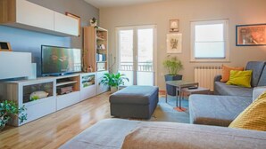 Una sala de estar moderna con televisor de pantalla plana, estanterías empotradas, un sofá gris y un comedor con una pequeña mesa y sillas.