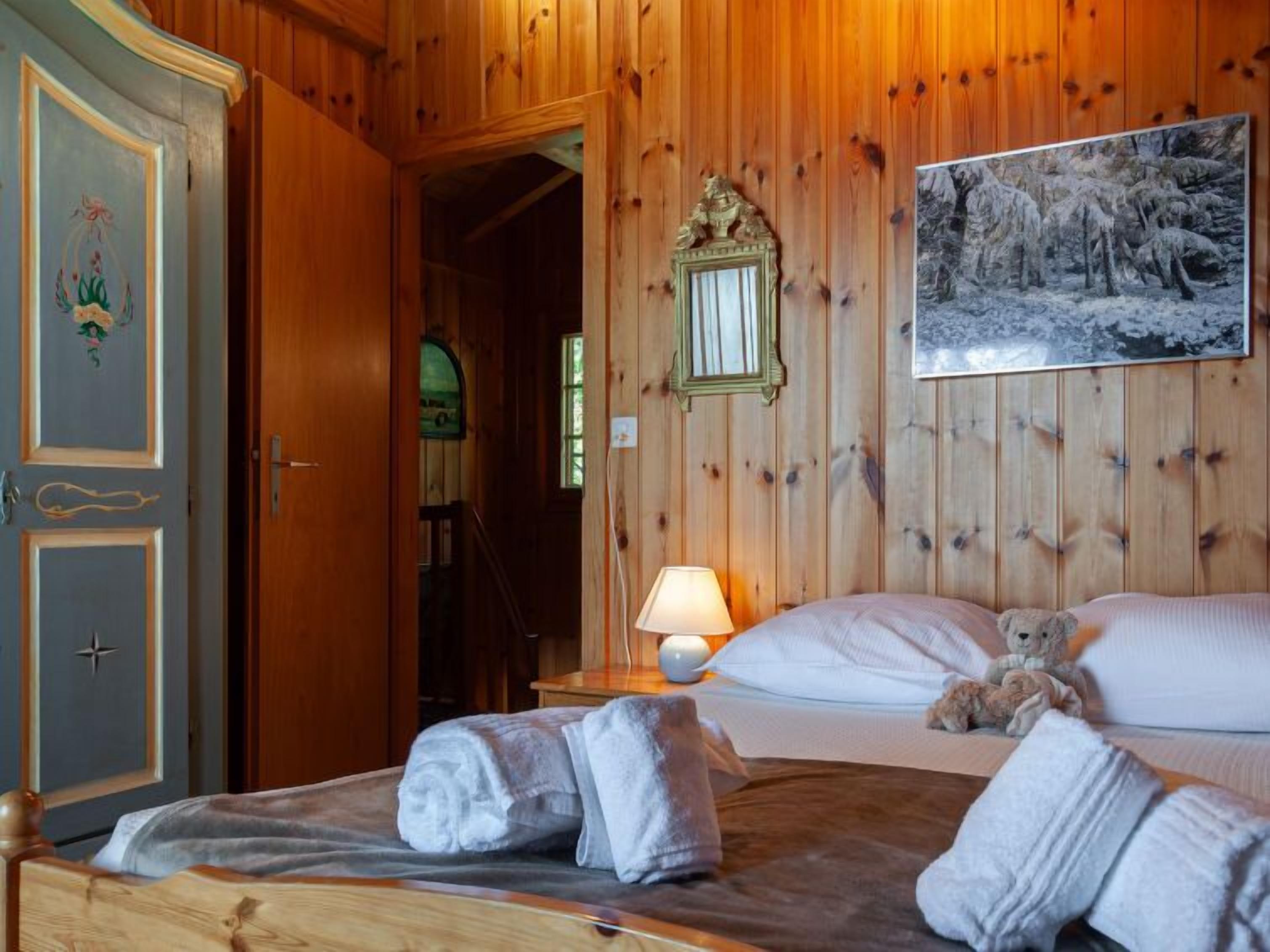 Ein Zimmer mit Holzwänden, einem Bett, einem Nachttisch mit Lampe, einem Teddybären und einem Gemälde von einem verschneiten Wald.