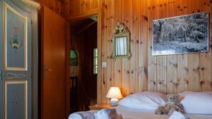 Een kamer met houten lambrisering, een bed, een nachtkastje met lamp, een teddybeer en een schilderij van een besneeuwd bos.
