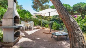 Eine Steinküche im Freien mit Pizzaofen, umgeben von einer Terrasse mit Tischen und Stühlen, unter einem großen Baum.