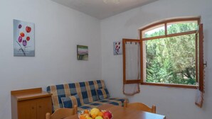 Ein Zimmer mit einem Holzesstisch, einem blau-orange karierten Sofa, einem Holzschrank und einem Fenster mit Blick auf Grünflächen.