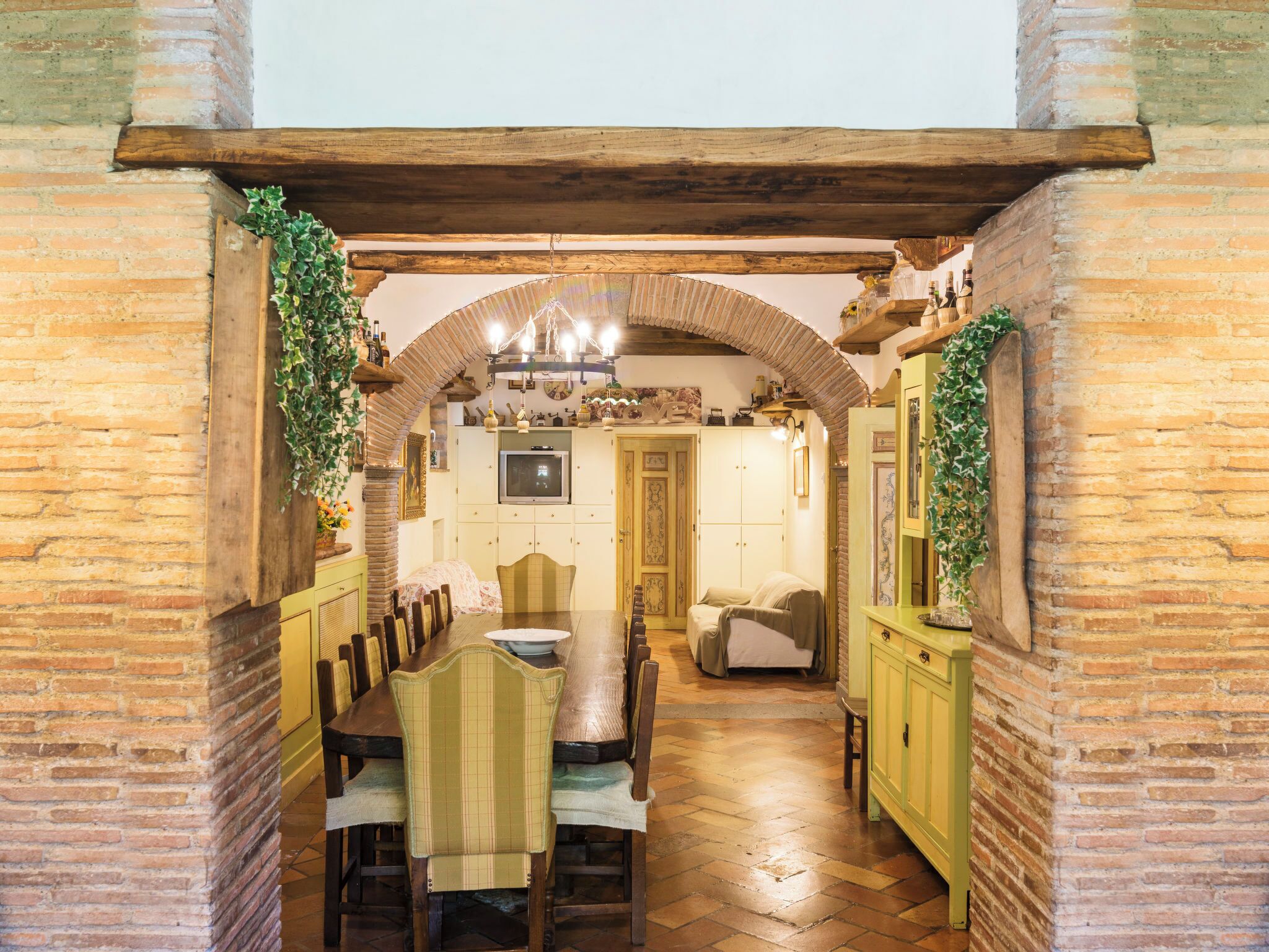 Zona pranzo con tavolo e sedie in legno, televisione sopra il tavolo e cucina sullo sfondo.