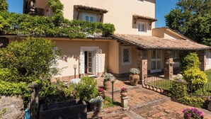 Un bâtiment à deux étages, doté d’un toit recouvert de tuiles, d’un balcon orné de plantes et d’un chemin en briques menant à l’entrée.