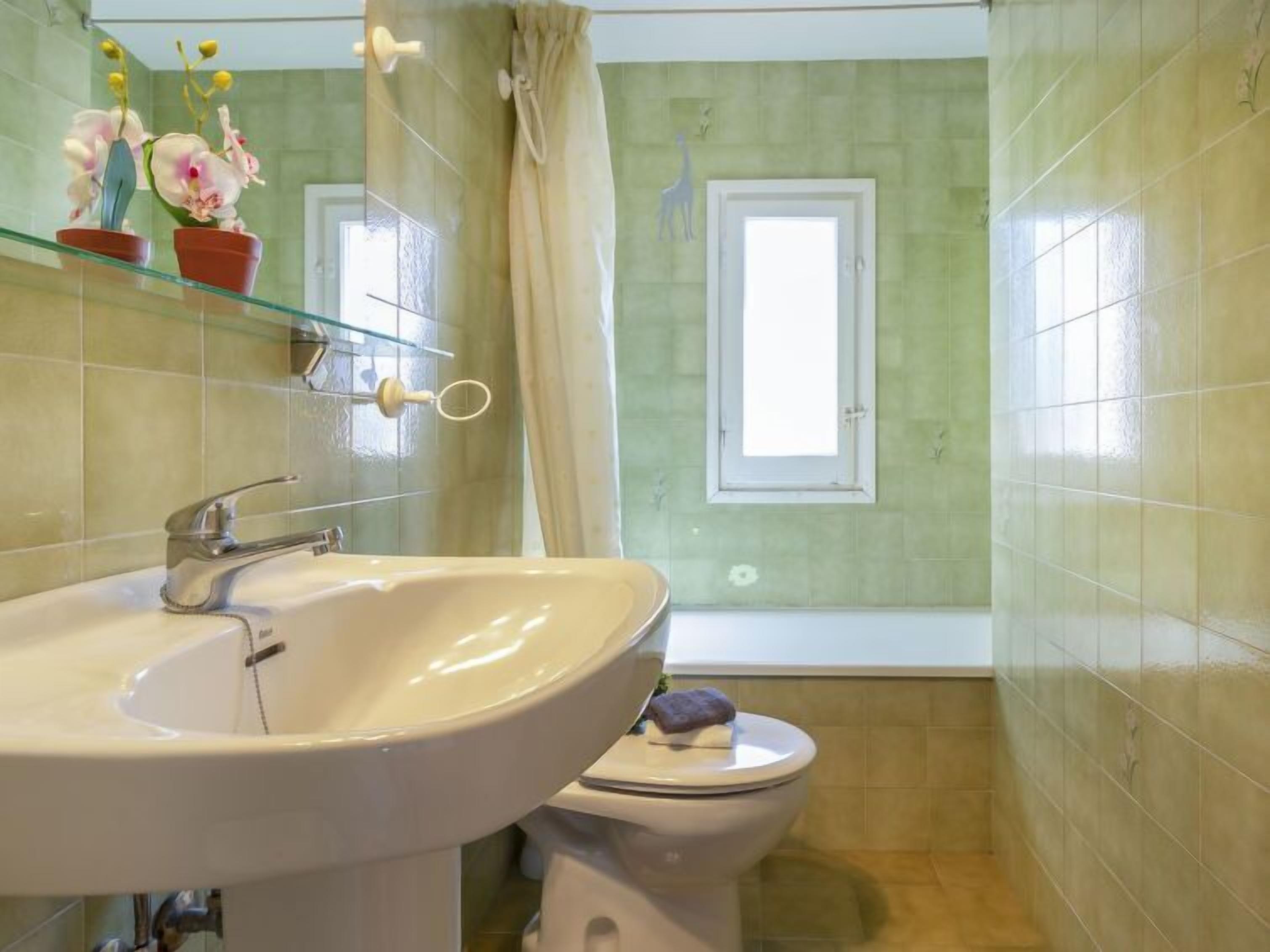 Ein Badezimmer mit einem weißen Waschbecken, einer Toilette, einem Glastischchen mit Blumen, einem Fenster und einer Duschvorhang.