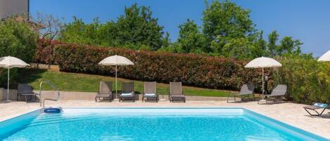 A clear blue swimming pool with lounge chairs and umbrellas, surrounded by greenery and a hedge.