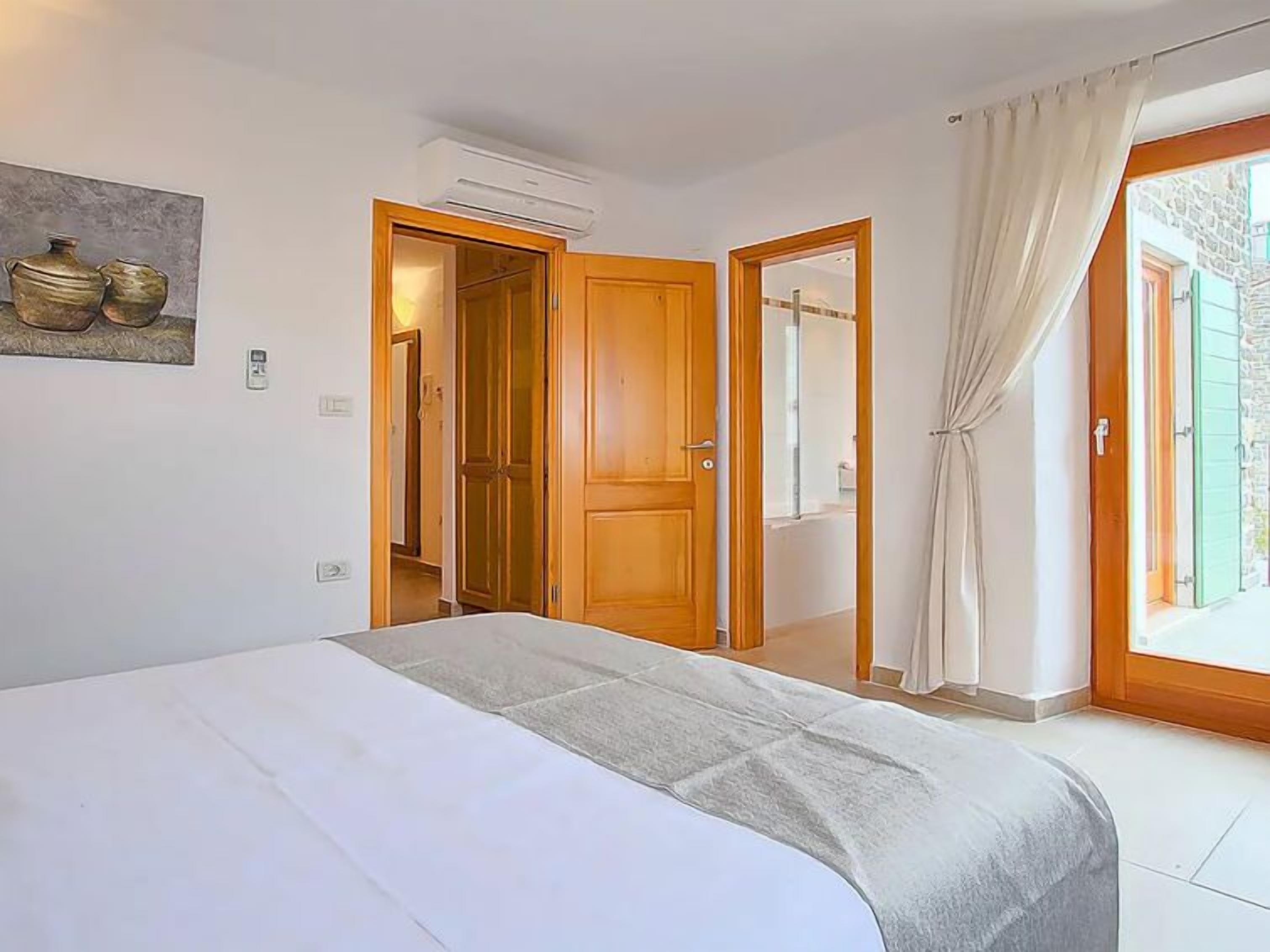 A bedroom with a bed, a wooden door, an air conditioning unit, and a view through a window with curtains.