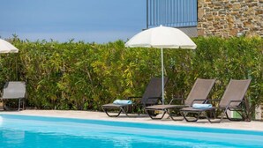 Uma área de piscina com espreguiçadeiras e guarda-sóis, cercada por vegetação e um muro de pedra.