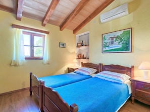 A room with two wooden beds, a window with curtains, a painting on the wall, and a wooden nightstand with a lamp.