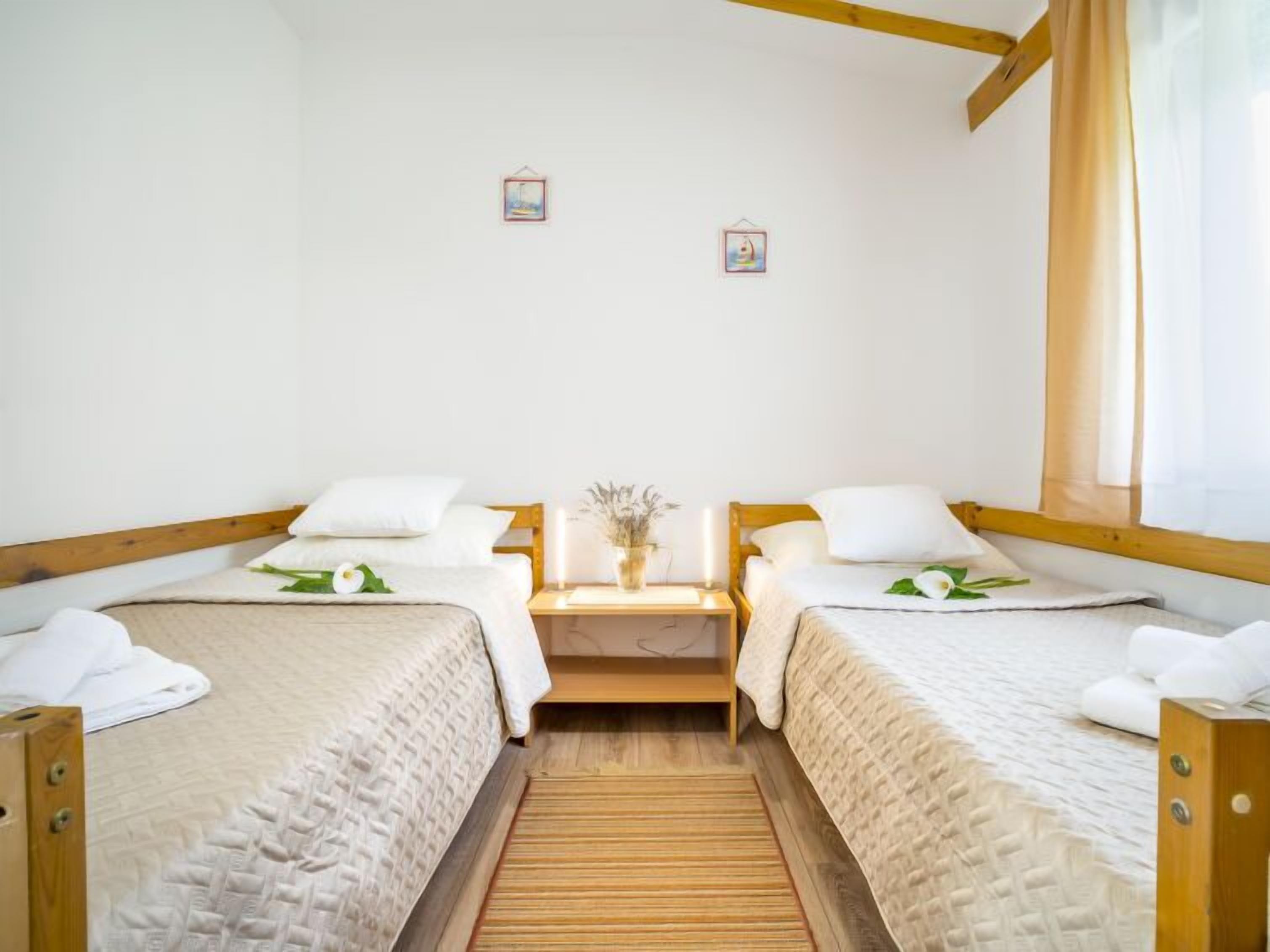 A room with two single beds, wooden headboards, white bedding, a small table with a vase, and a window with curtains.