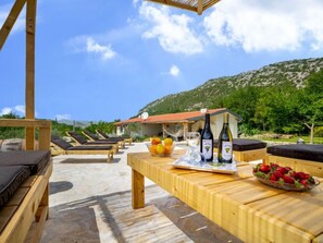 A wooden table with wine bottles, fruit, and glasses, set against a backdrop of a mountain and a clear sky. - Vacation home Jozo by Interhome (Omiš)