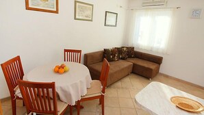 A dining area with a round table, chairs, and a small round table with a woven placemat. There is a brown sofa with patterned cushions against a wall with framed pictures. An air conditioning unit is mounted on the wall above a window with sheer curtains. - Parunov by Interhome (Šolta/Rogac)