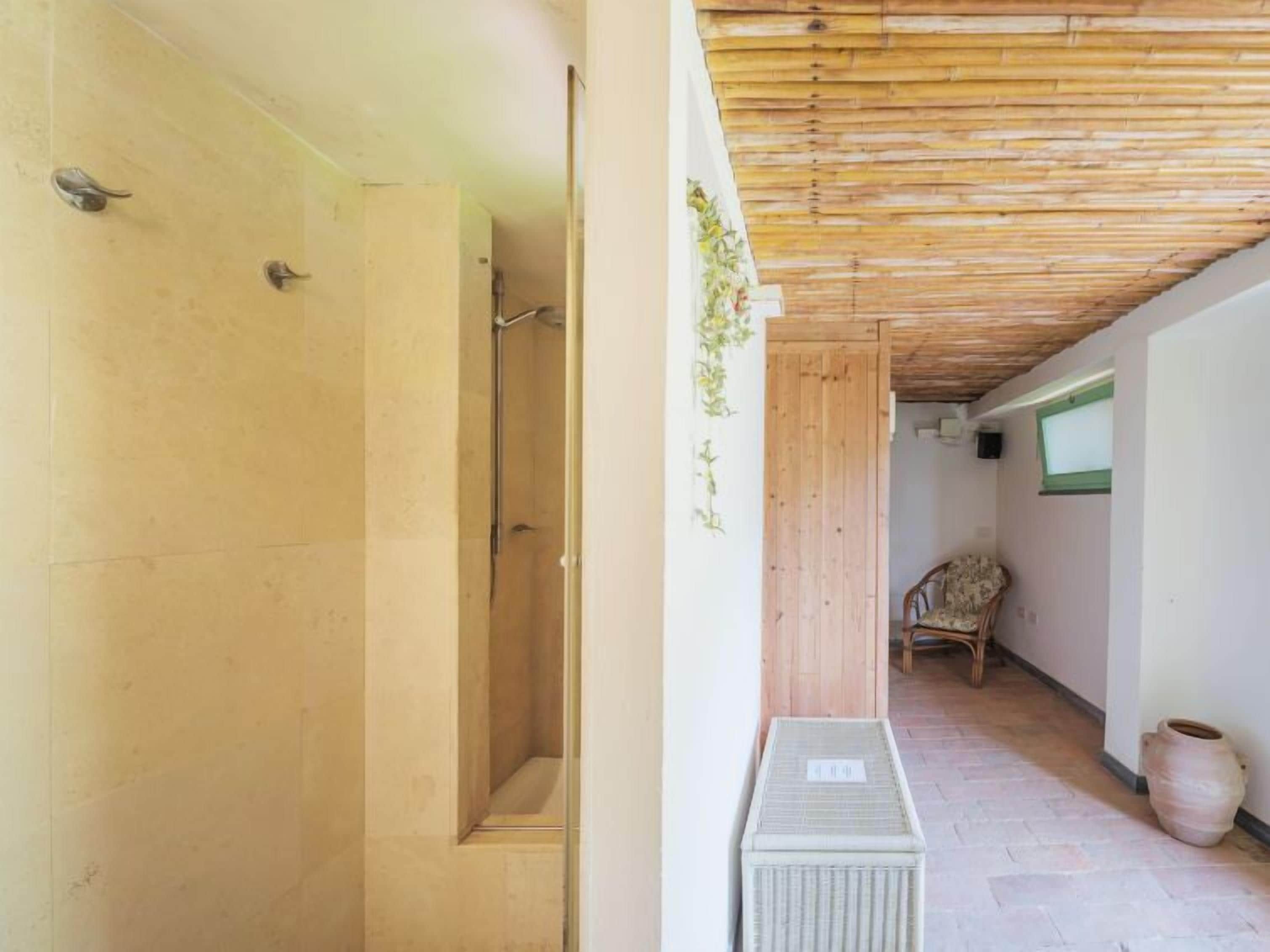 A hallway with a wooden ceiling, a shower area, a wicker chair, and a tiled floor.