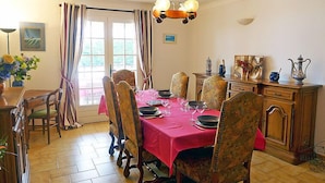 Comedor con una mesa dispuesta para seis personas, rodeada por seis sillas. Hay una araña colgante sobre la mesa, y una ventana con cortinas a la izquierda.