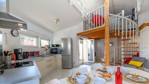 Une cuisine moderne dotée d’un îlot central, un coin repas avec une table mise pour le repas, et une chambre en mezzanine avec un lit rouge.