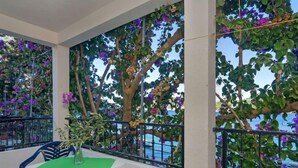 Un balcón con mesa y sillas, rodeado de abundante vegetación y flores moradas.