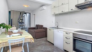 Cocina compacta con muebles blancos, lavavajillas y horno. Cuenta con un comedor con mesa y sillas, y un sofá marrón.