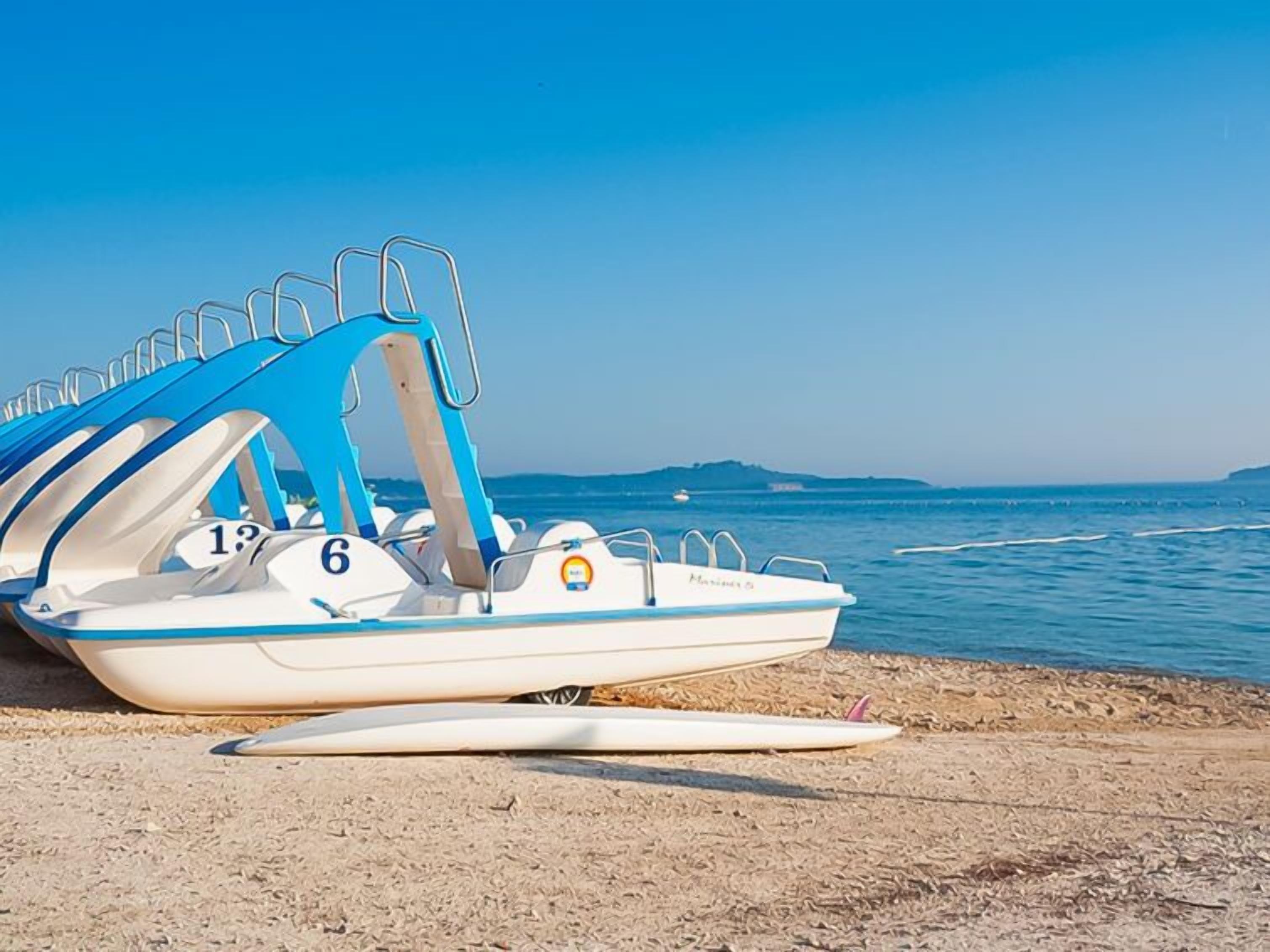 A row of small, white boats with blue accents, numbered 1 to 6, docked on a sandy beach with the sea in the background.