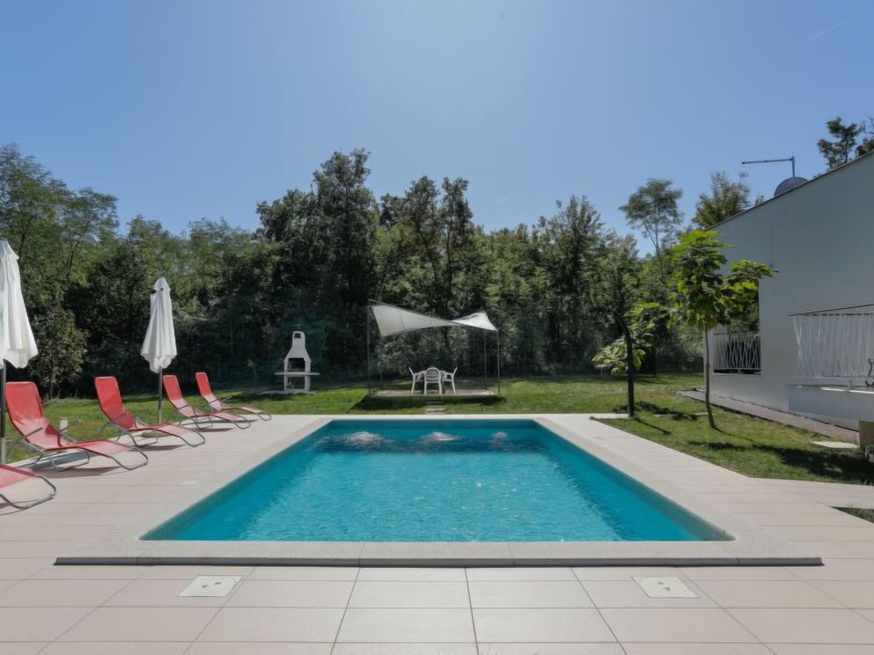 En rektangulär pool med en kakelbeklädd poolkant, omgiven av röda solstolar och vita parasoller.
