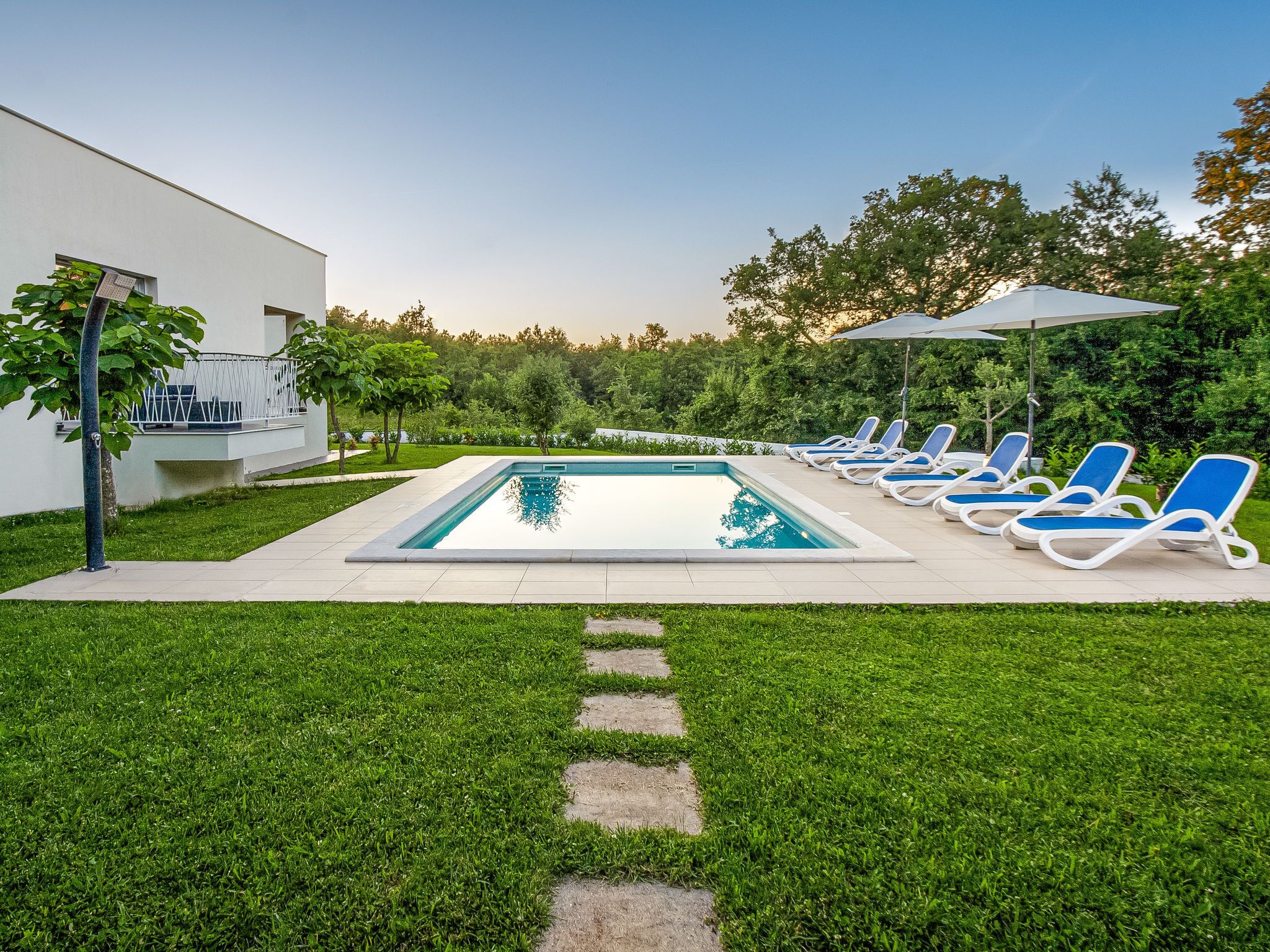 Une piscine rectangulaire à l’eau d’un bleu limpide, entourée d’une terrasse dallée. Plusieurs chaises longues avec des coussins bleus sont disposées autour de la piscine, et un grand parasol offre de l’ombre. L’espace piscine est bordé d’une pelouse bien entretenue, avec un chemin de pierre menant à la piscine.