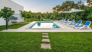 Ein rechteckiger Swimmingpool mit glasklarer, blauer Wasserfläche, umgeben von einer gepflasterten Liegefläche. Mehrere Liegestühle mit blauen Kissen sind um den Pool verteilt, und ein großer Sonnenschirm spendet Schatten. Das Pool-Areal wird von einer gepflegten Rasenfläche mit einem steinernen Pfad zum Pool gesäumt.