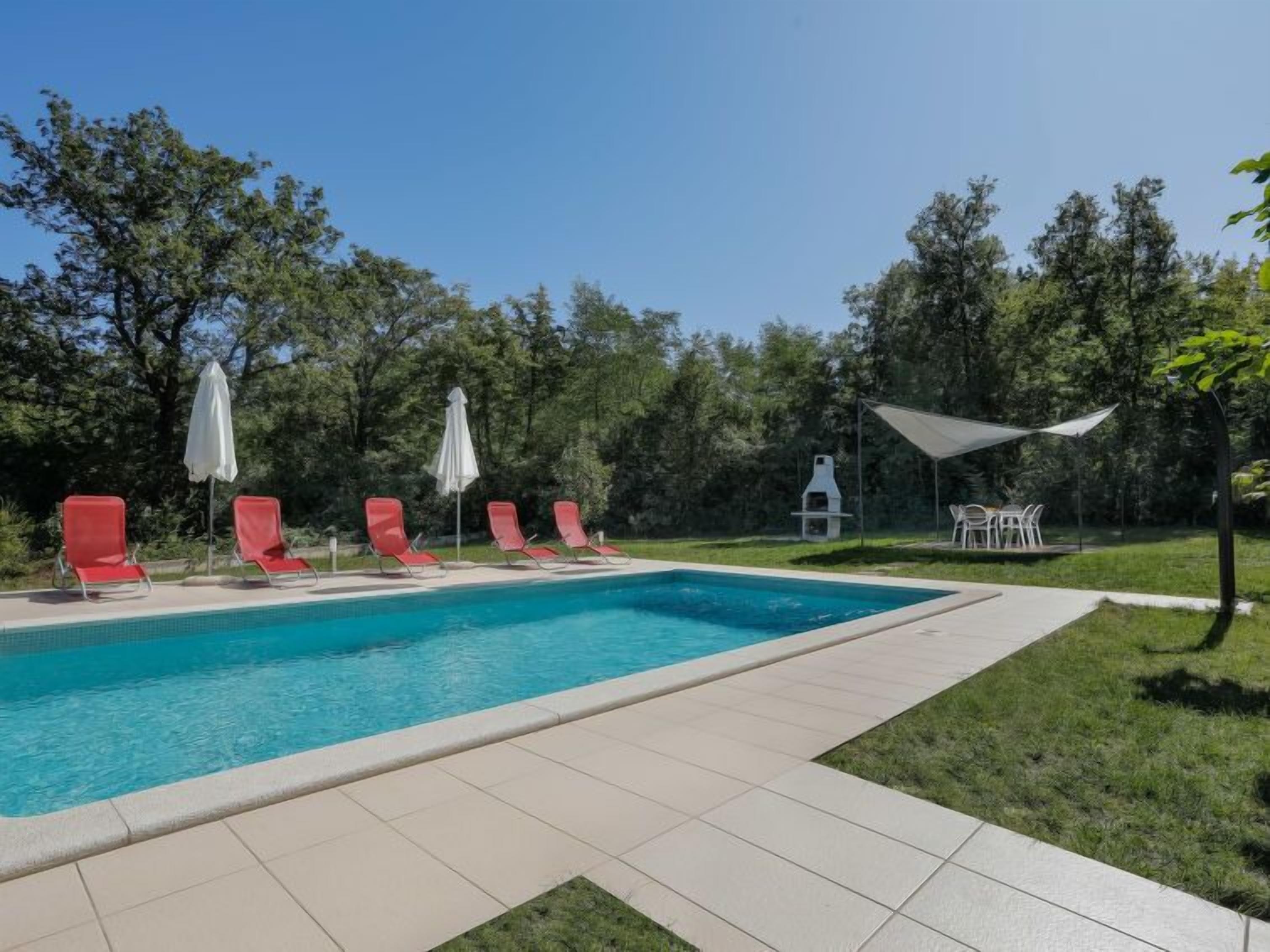 A rectangular swimming pool with a tiled edge, surrounded by a grassy area with red lounge chairs and white umbrellas.