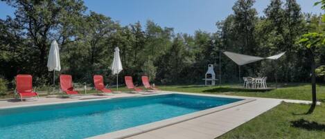 A rectangular swimming pool with a tiled edge, surrounded by a grassy area with red lounge chairs and white umbrellas.