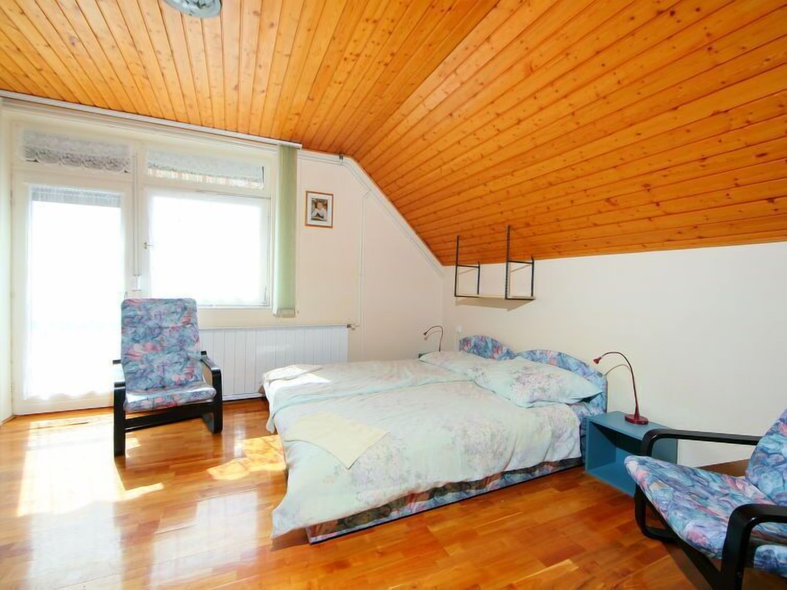 A bedroom with a wooden ceiling, a bed with floral bedding, a chair, and a bedside table.