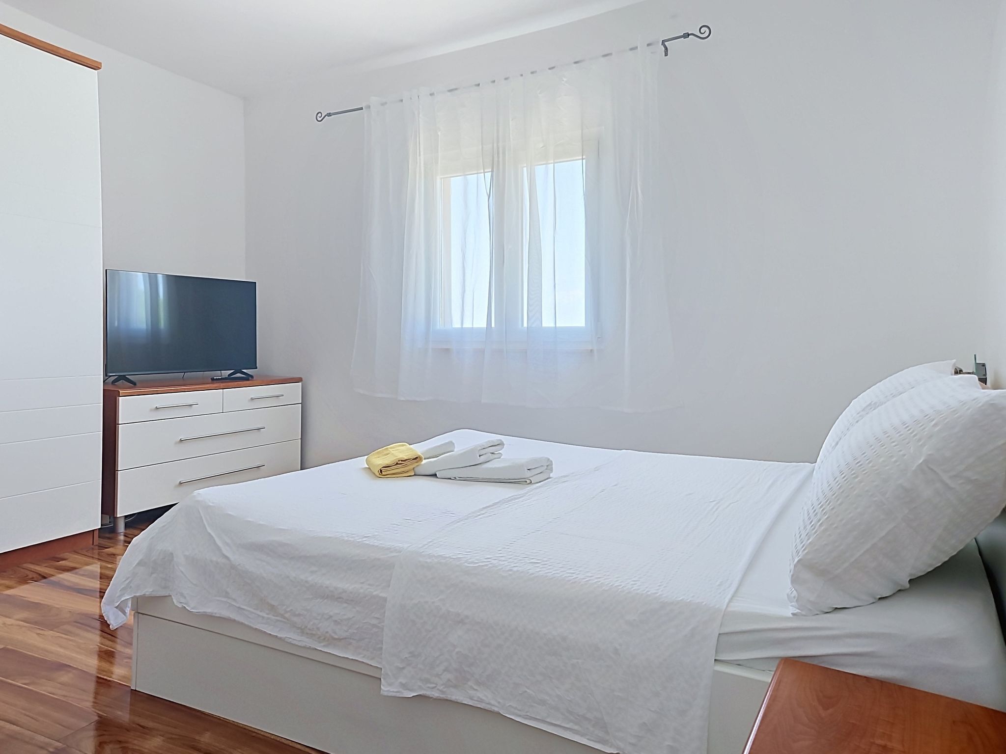 Een slaapkamer met een bed, een televisie op een houten standaard, een raam met dunne gordijnen en een houten vloer.