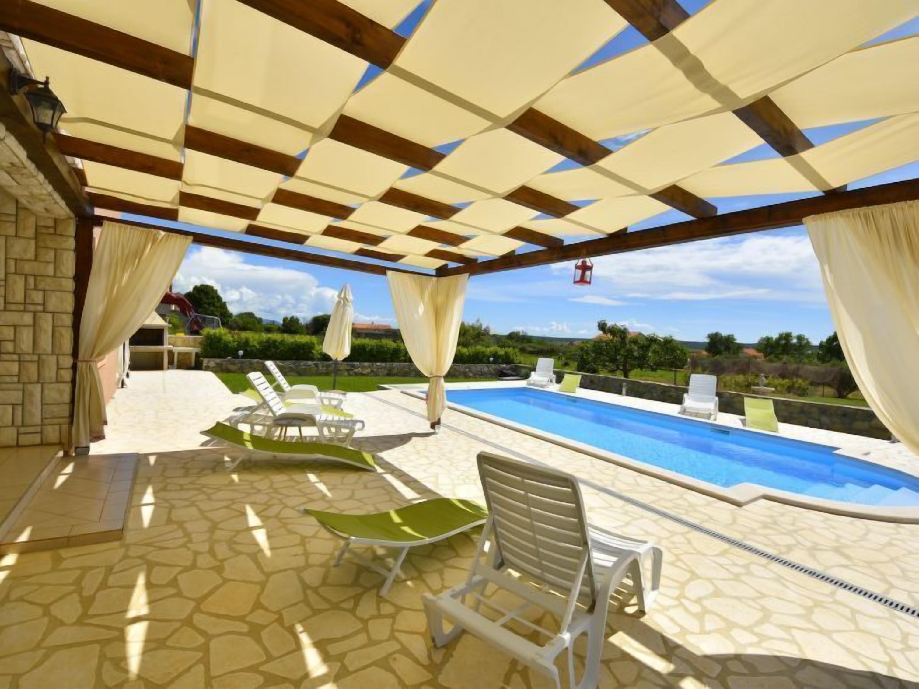A covered outdoor swimming pool area with lounge chairs and a stone-tiled floor.