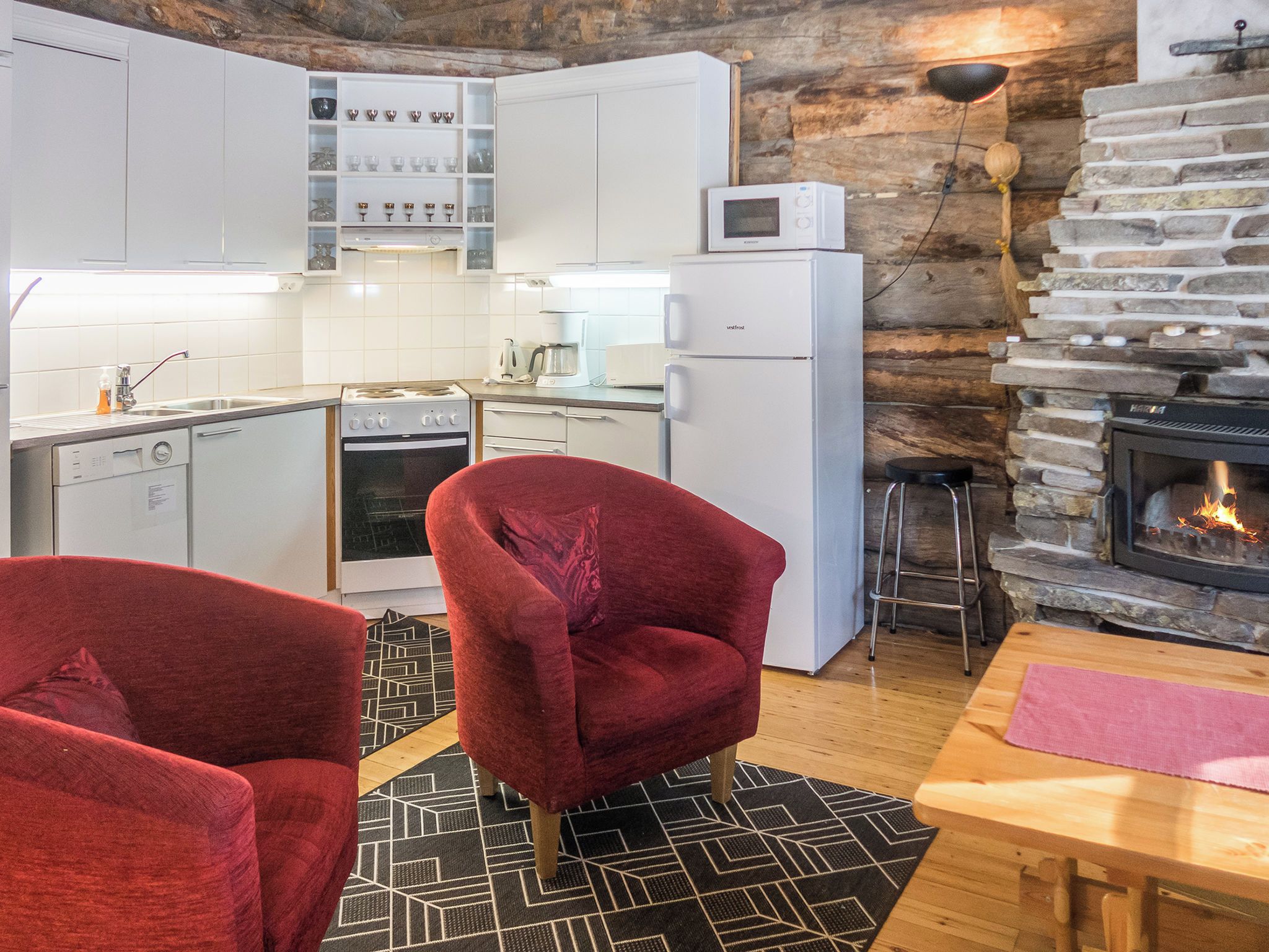 A compact kitchen with white cabinets, a stove, and a microwave. There are two red chairs and a wooden table with a rug. A stone fireplace is visible in the background.