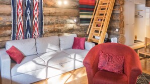A log cabin interior with a sofa, red armchair, wooden ladder, and colorful blankets.