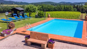 A rectangular swimming pool with a tiled edge, surrounded by a brick patio. There are blue lounge chairs and a small wooden structure in the background.