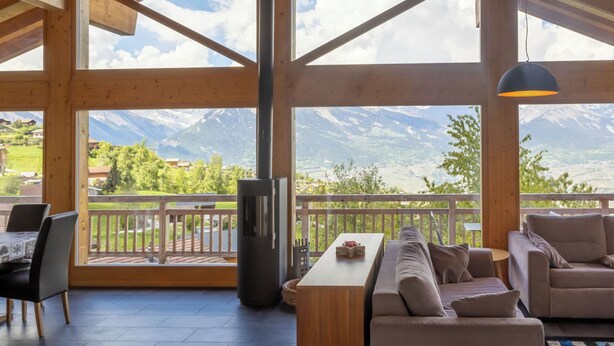 A spacious room with a wooden ceiling, large windows, a sofa, a table, and a hanging light fixture. - Vacation home D'Arby by Interhome (Nendaz)