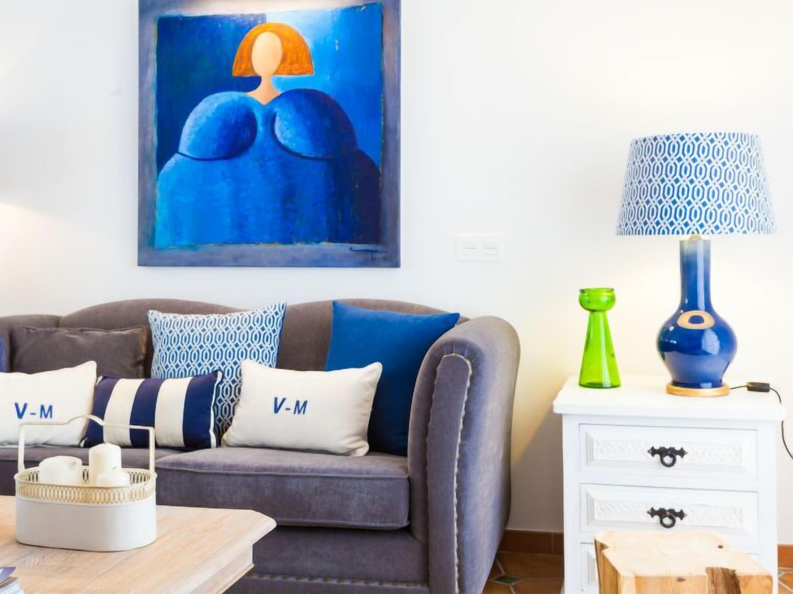 A living room with a grey sofa, blue and white patterned cushions, a blue and white patterned lamp, and a painting on the wall.