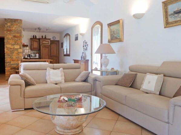 Una sala de estar con dos sofás color beige, una mesa de centro de vidrio y una zona de cocina al fondo.