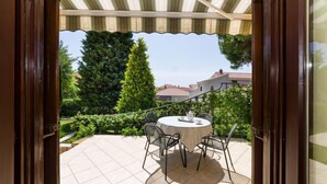 An outdoor patio with a round table and chairs, a striped awning, and a view of a residential area with trees and houses.