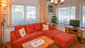 Een gezellige woonkamer met een rode hoekbank, een houten salontafel, een televisie op een houten meubel en een groot raam met gordijnen.