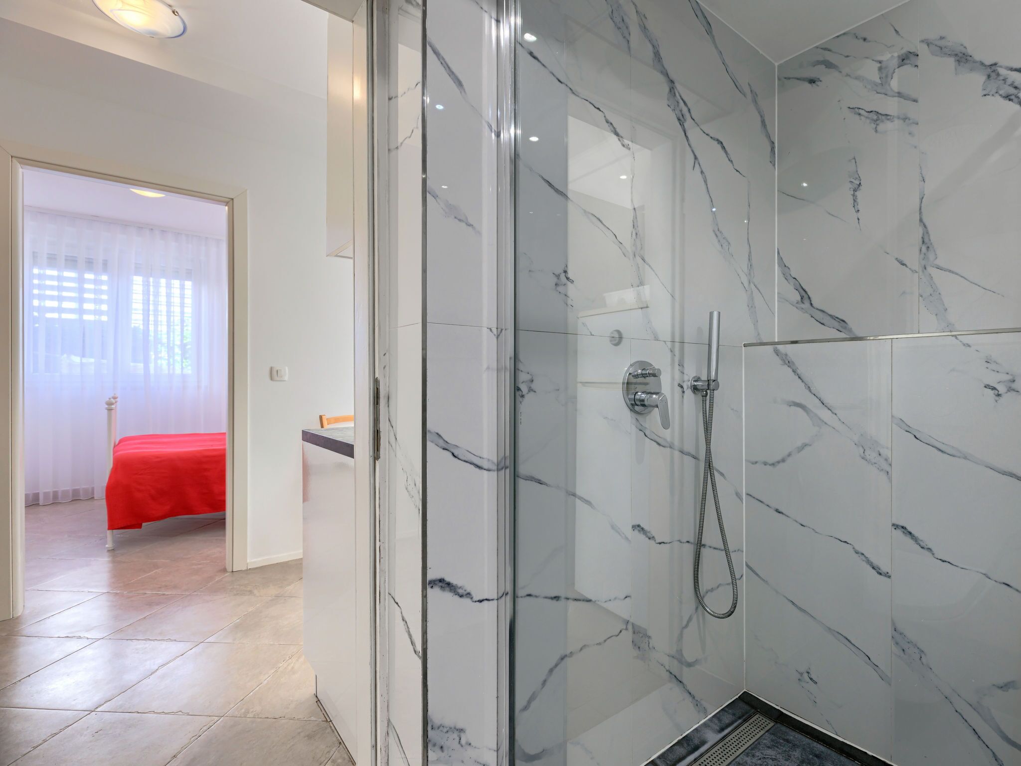 A modern bathroom with a glass-enclosed shower, marble walls, and a bed in the adjacent room.