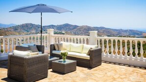 Eine Terrasse mit Korbmöbeln, einem Tisch und einem Sonnenschirm. Von der Terrasse hat man einen Blick auf eine gebirgige Landschaft.