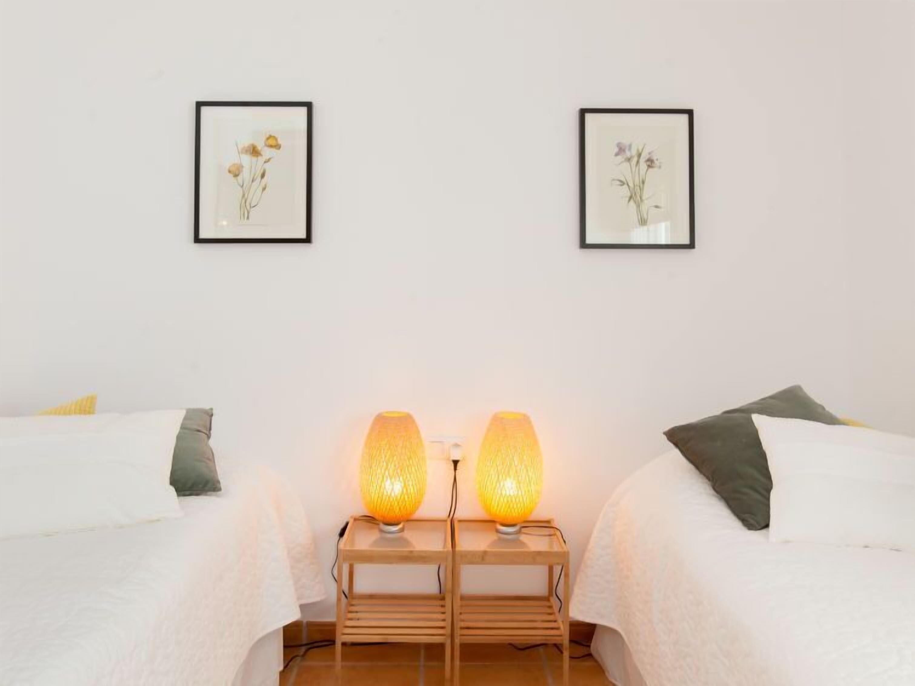 Ein Hotelzimmer mit zwei Betten, jeweils mit einem Kopfkissen, einem Nachttisch mit Lampe und zwei gerahmten Blumenbildern an der Wand.