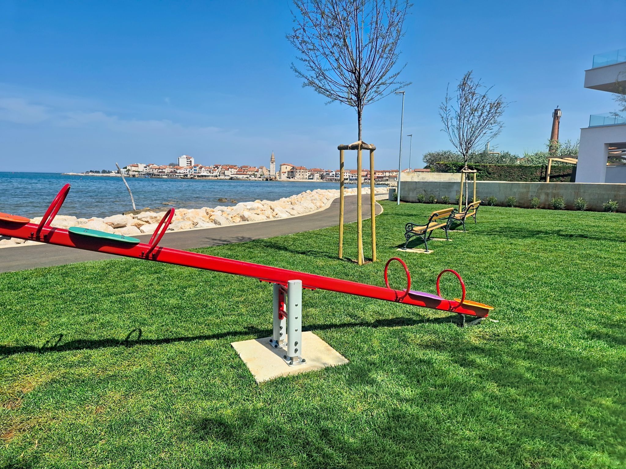 A park with seesaws, benches, and a view of the sea and buildings in the background.